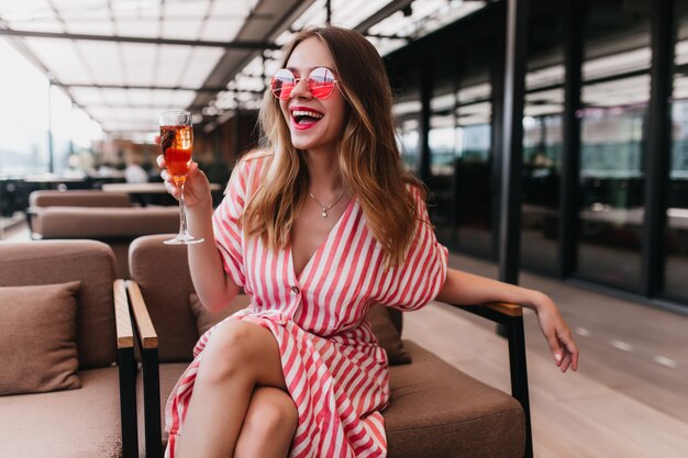 Dichosa chica rubia pasando el día de verano en el restaurante. Feliz mujer europea viste vestido de rayas bebiendo champán y sonriendo.