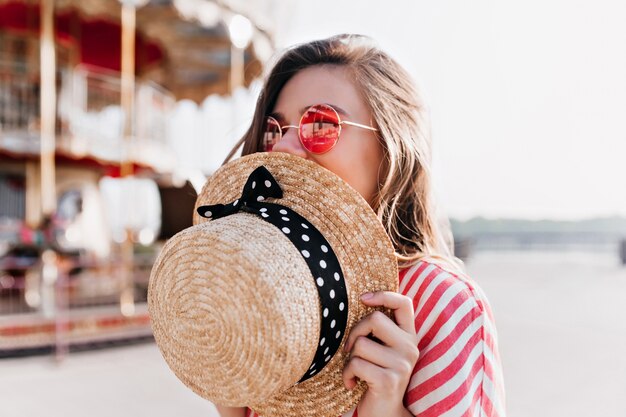Dichosa chica rubia que cubre la cara con sombrero de paja mientras posa en día de verano. Foto al aire libre de mujer joven feliz con gafas de sol rosas descansando en el parque de atracciones.