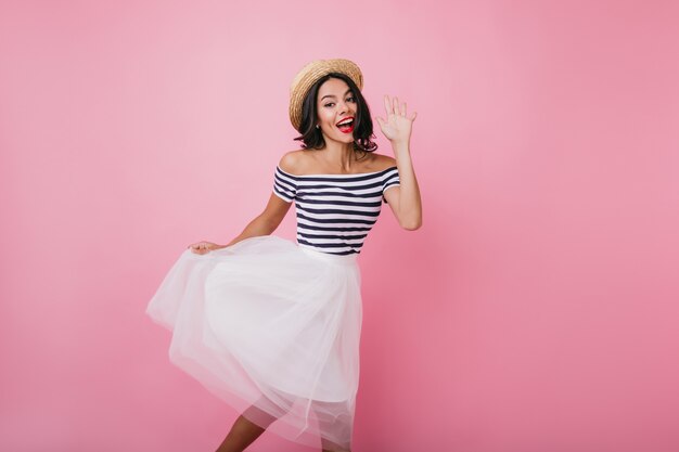 Dichosa mujer bronceada agitando la mano mientras baila. Retrato interior de niña morena complacida viste elegante falda exuberante.