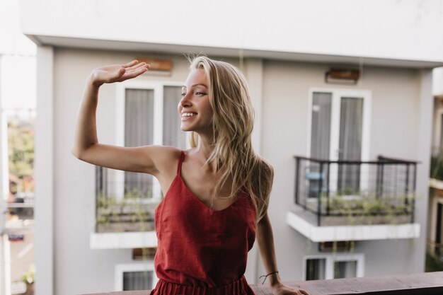 Dichosa mujer bronceada en pijama agitando la mano con una sonrisa. Increíble modelo de mujer caucásica de pie en el balcón.