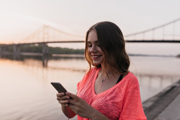 Dichosa mujer europea usando su teléfono en el río. Chica romántica caminando cerca del lago y mensaje de texto.