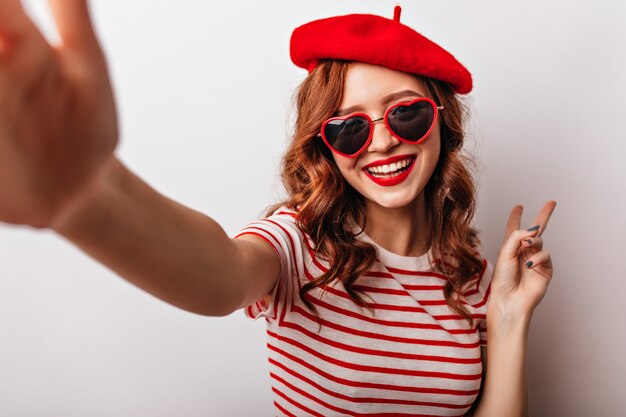 Dichosa mujer francesa en gafas de sol haciendo selfie. Entusiasta chica pelirroja rizada posando en la pared blanca.