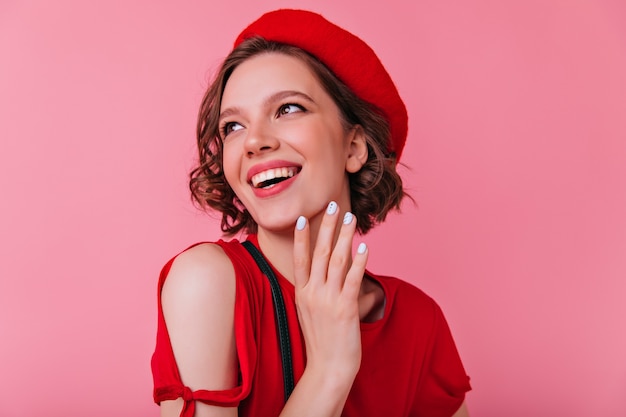 Dichosa mujer francesa con manicura blanca riendo. Filmación en interiores de alegre chica rizada en boina roja mirando a otro lado con una sonrisa.