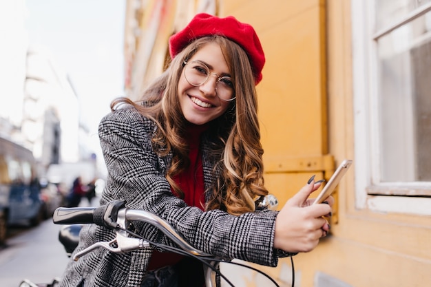 Dichosa niña caucásica lleva chaqueta de tweed leyendo un mensaje telefónico en el fondo de la ciudad