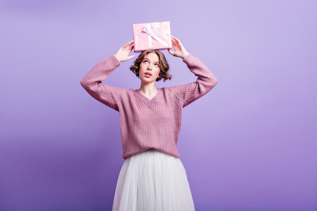 Foto gratuita dichoso modelo femenino europeo con regalo de cumpleaños sobre la cabeza y sonriendo. chica fascinante con corte de pelo corto posando con regalo en pared coloful.