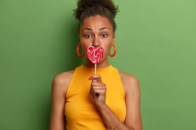 Foto gratuita dieta de azúcar, concepto de comida poco saludable. encantadora mujer adulta de piel oscura cubre la boca con piruleta, disfruta de un postre dulce, disfruta de las vacaciones de verano, viste una camiseta amarilla, aislada en la pared verde.