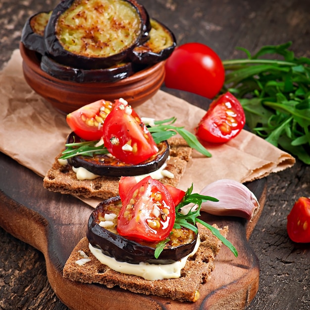 Dieta vegetariana Sandwiches de pan crujiente con queso crema de ajo, berenjenas asadas, rúcula y tomates cherry sobre superficie de madera vieja