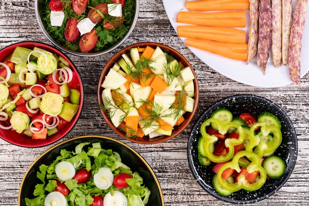 Diferentes ensaladas de verduras en diferentes tazones sobre superficie de madera blanca frijoles pimiento zanahoria cebolla lechuga vista superior