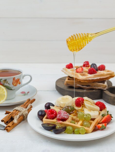 Diferentes frutas con gofres en un plato con miel, canela, taza de té