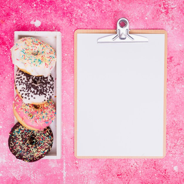Diferentes tipos de donuts en una caja rectangular blanca cerca del portapapeles con papel blanco sobre fondo rosa