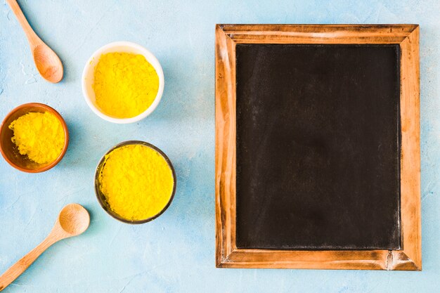 Diferentes tipos de tazones con polvo de color amarillo holi y una cuchara cerca de la pizarra de madera en blanco