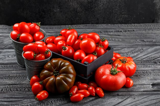 Diferentes tomates en caja de madera, mini cubos en madera gris y pared oscura, vista de ángulo alto.