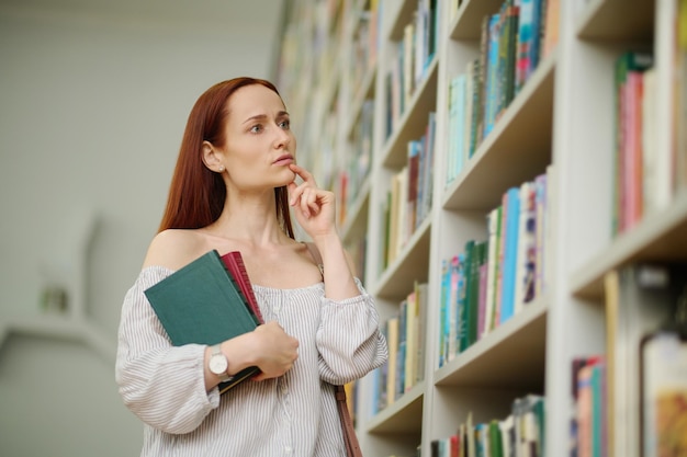 Dificultad de elección. Mujer joven de pensamiento serio con el pelo largo y rojo de pie con libros en la mano cerca de la estantería tocando la barbilla con el dedo en la biblioteca