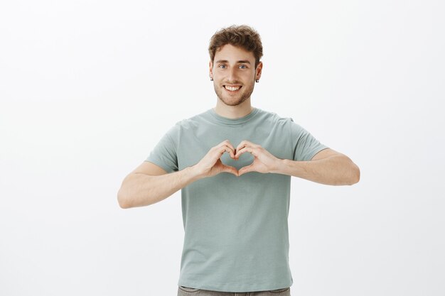 Difunde amor, no guerra. Retrato de feliz chico positivo de aspecto amistoso con aretes que muestra un gesto de corazón sobre el pecho y una amplia sonrisa mientras expresa afecto o emociones románticas sobre una pared gris