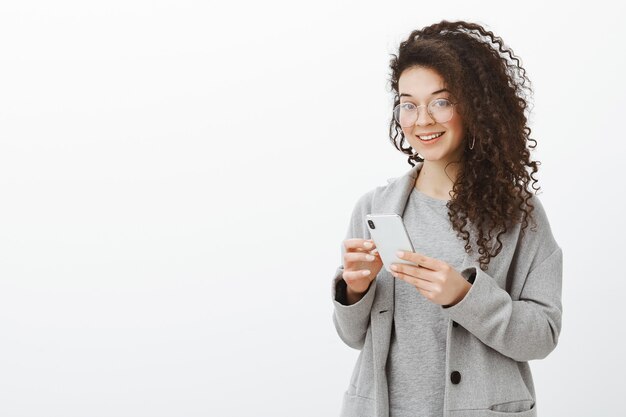 Dime tu numero de telefono. Retrato de mujer segura de buen aspecto elegante en gafas de moda y abrigo gris, sosteniendo el teléfono inteligente y mirando con interés y alegría