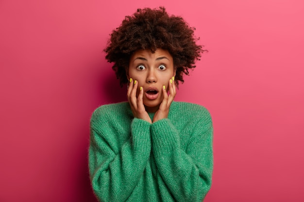 ¡Dios mío, es horrible! La mujer sin palabras asustada mantiene ambas manos en las mejillas, mira fijamente con los ojos abiertos, teme a algo aterrador, se encuentra insegura, vestida con ropa verde, aislada en la pared rosa