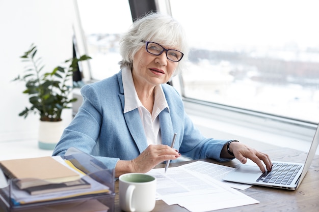 Director ejecutivo femenino serio atractivo de la edad madura que se sienta en su oficina con el ordenador portátil, mecanografía y firma papeles en el escritorio, que tiene mirada confiada. Concepto de personas, envejecimiento, trabajo y carrera