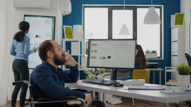 Discapacitado inválido empresario discapacitado sentado en silla de ruedas trabajando en la computadora con estadísticas financieras contestando en el teléfono