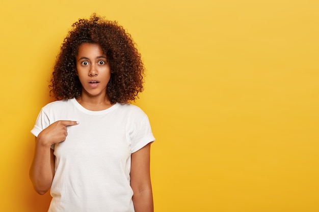 ¿Disculpe? Dama afroamericana indignada se siente perpleja, se señala a sí misma, sorprendida por palabras ofensivas, se ve confusa, usa ropa blanca, se para contra la pared amarilla, espacio en blanco a un lado