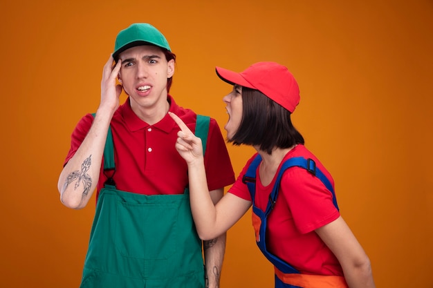 Discutiendo a una pareja joven en uniforme de trabajador de la construcción y gorra estresada chico tocando la cabeza mirando hacia abajo chica enojada de pie en la vista de perfil mirando y gritando a chico levantando el dedo