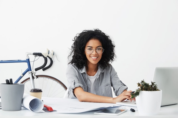 Diseñador de estudiante femenino sonriente sentado en su lugar de trabajo rodeado de aparatos