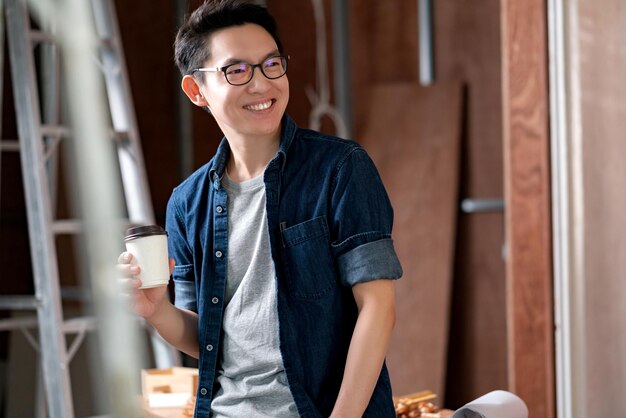 Diseñador de interiores de lentes atractivos asiáticos que trabaja en el progreso de la construcción de la renovación del sitio de la casa con una sonrisa y confianza en el escritorio con una herramienta de impresión y dibujo azul