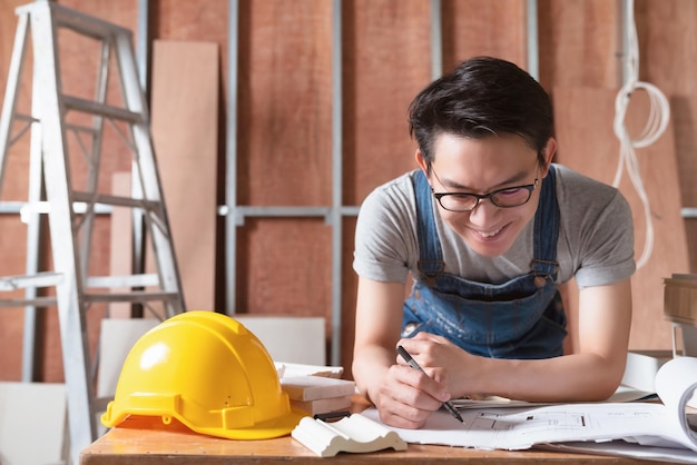 Diseñador de interiores de lentes atractivos asiáticos que trabaja en el progreso de la construcción de la renovación del sitio de la casa con una sonrisa y confianza en el escritorio con una herramienta de impresión y dibujo azul