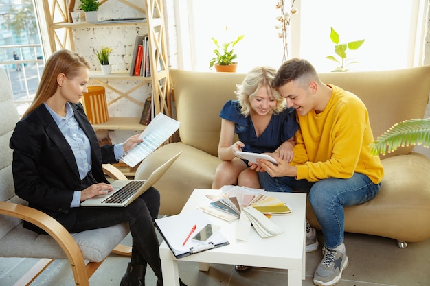 Diseñador de interiores que trabaja con pareja joven. Encantadora familia y diseñador o arquitecto profesional discutiendo el concepto del futuro interior, trabajando con paleta de colores, dibujos de habitaciones en la oficina moderna.