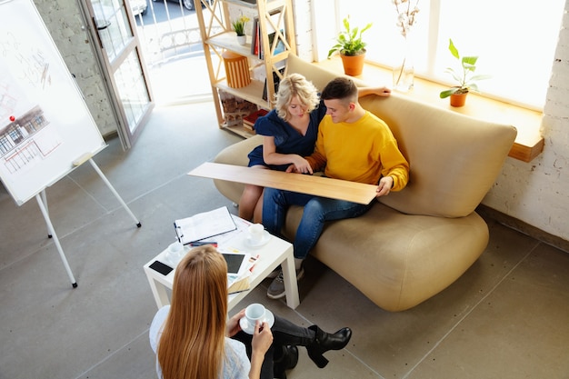 Diseñador de interiores que trabaja con pareja joven. encantadora familia y diseñador o arquitecto profesional discutiendo el concepto del futuro interior, trabajando con paleta de colores, dibujos de habitaciones en la oficina moderna.