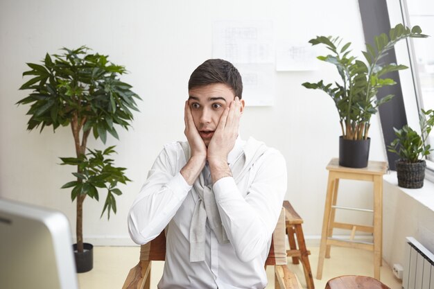 Diseñador masculino desconcertado gesticulando presa del pánico, mirando la pantalla de la computadora, con una mirada de asombro y preocupación porque no puede terminar el dibujo del plan de construcción a tiempo. Plazo y estrés en el trabajo