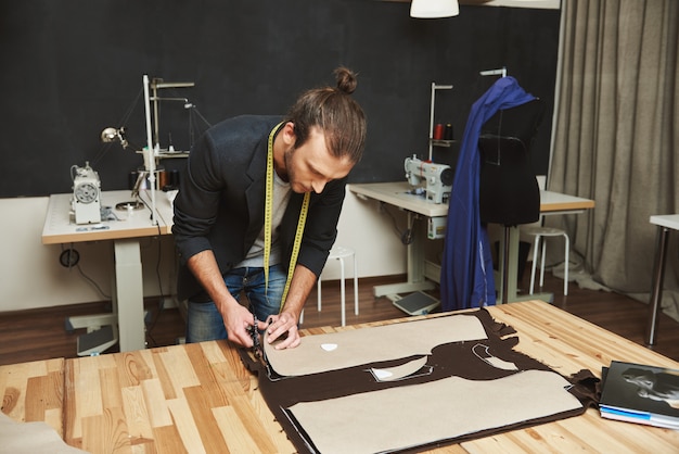 Diseñador de moda masculino caucásico guapo creativo oscuro maduro en traje negro cortando partes de ropa de tela con tijeras, pasando la noche en el estudio.