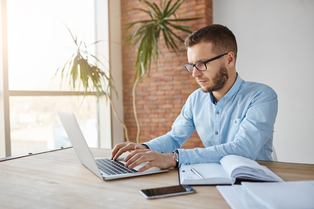 Foto gratuita diseñador web freelance masculino maduro sentado en un espacio de trabajo conjunto, trabajando en una computadora portátil, escribiendo tareas en el cuaderno