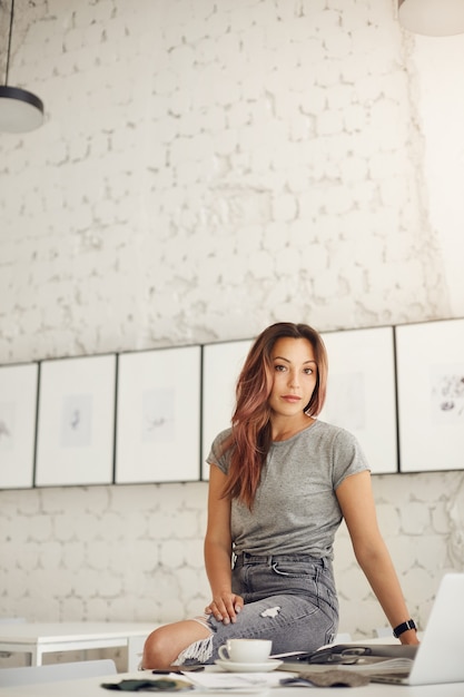 Diseñadora de moda femenina que trabaja en la computadora portátil tomando café en un luminoso espacio de estudio. Joven talento abriéndose camino en la vida. Mirando a la cámara.