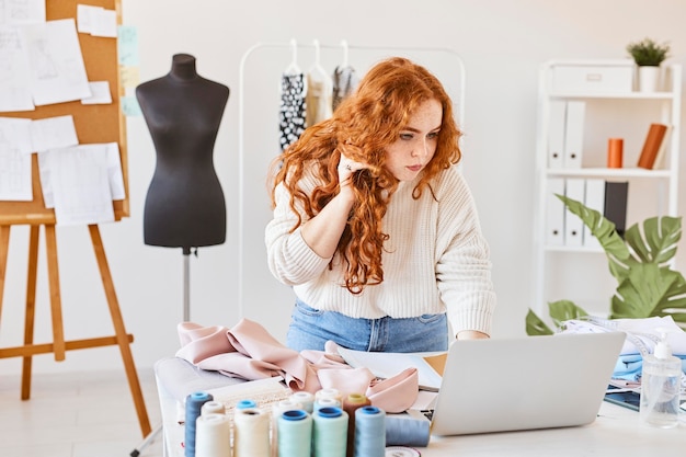 Diseñadora de moda femenina trabajando en atelier con laptop y hablando por teléfono inteligente