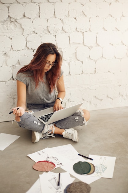 Foto gratuita diseñadora de moda femenina trabajando en una computadora portátil en su estudio comprobando telas y bocetos sentados en el piso. industria creativa.