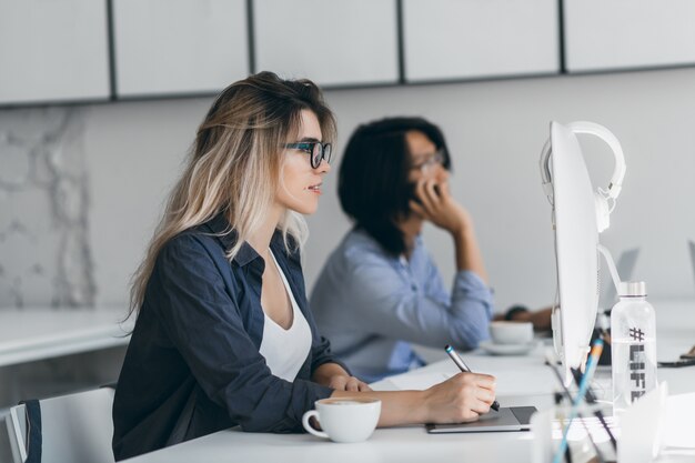 Diseñadora web independiente inspirada que usa tableta y lápiz, mirando la pantalla mientras su amiga habla por teléfono. Estudiante asiático sosteniendo el teléfono inteligente y escribiendo en el teclado, sentado al lado de la chica rubia.