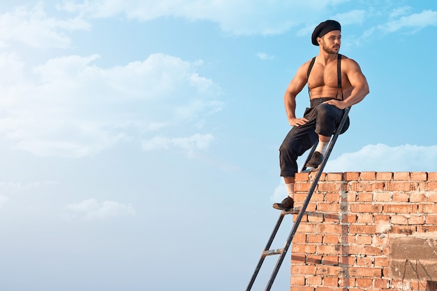 Disfrutando al aire libre. Disparo horizontal de un trabajador de la construcción sin camisa sexy sentado en una escalera mirando a otro lado con alegría cielos azules en el fondo