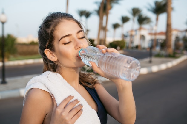 Disfrutando de la mañana soleada, bebiendo agua de la botella con los ojos cerrados de una joven bastante deportiva en el entrenamiento exterior. Entrenamiento, ciudad tropical, relax, estilo de vida saludable