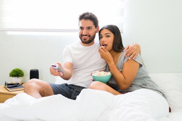 Disfrutando de una noche de cine. Un joven feliz poniendo una serie de televisión y abrazándose con su novia en la cama mientras come palomitas de maíz