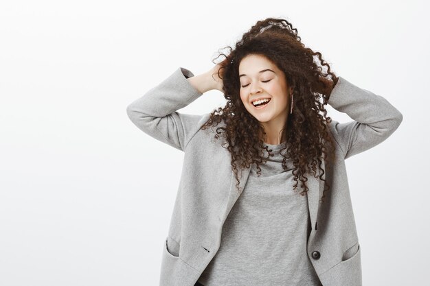 Disfrutando la vida al máximo. feliz niña soñadora femenina con cabello rizado, cerrando los ojos, tocando la cabeza, sonriendo con alegría, expresando una actitud tierna y tímida