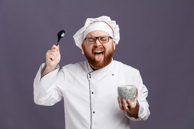 Disgustado joven chef con gafas uniformes y gorra sosteniendo un tazón que muestra una cuchara y lengua con los ojos cerrados aislados en un fondo morado
