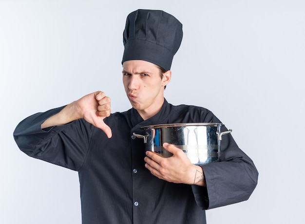 Disgustado joven cocinero de sexo masculino rubio en uniforme de chef y gorra sosteniendo la olla mirando a la cámara mostrando el pulgar hacia abajo aislado en la pared blanca