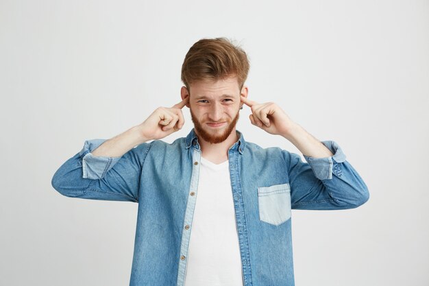 Disgustado joven guapo con barba frunciendo el ceño cerrando los oídos.