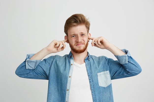 Disgustado joven guapo con barba frunciendo el ceño cerrando los oídos.