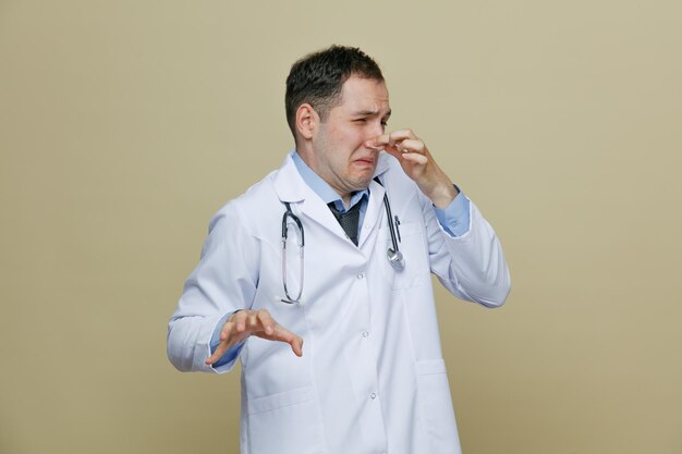Disgustado joven médico masculino con bata médica y estetoscopio alrededor del cuello manteniendo la mano en el aire mirando al costado haciendo un gesto de mal olor aislado en un fondo verde oliva