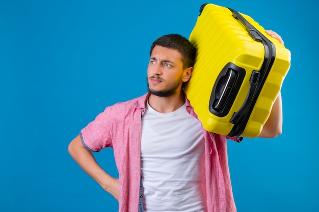 Disgustado joven viajero guapo chico sosteniendo la maleta mirando a un lado con el ceño fruncido de pie sobre fondo azul.