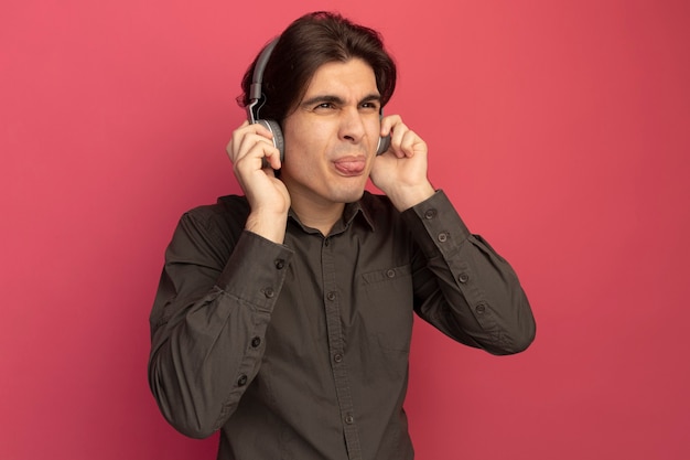 Disgustado mirando al lado chico guapo joven con camiseta negra con auriculares mostrando la lengua aislada en la pared rosa