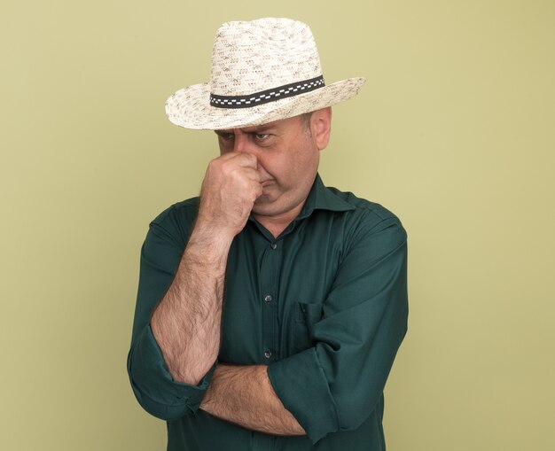 Disgustado mirando al lado hombre de mediana edad vestido con camiseta verde y sombrero agarró la nariz aislada en la pared verde oliva