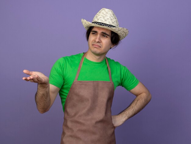 Disgustado mirando al lado joven jardinero en uniforme vistiendo puntos de sombrero de jardinería con la mano al lado