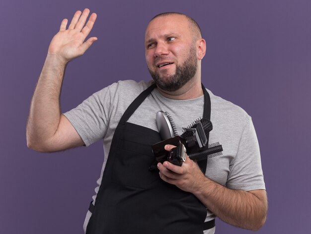 Disgustado mirando al peluquero masculino de mediana edad eslavo lateral en uniforme sosteniendo herramientas de peluquero levantando la mano aislada en la pared púrpura